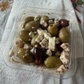 A small clear plastic container filled with green olives, feta cheese, and other ingredients in oil, sitting on a white cloth.