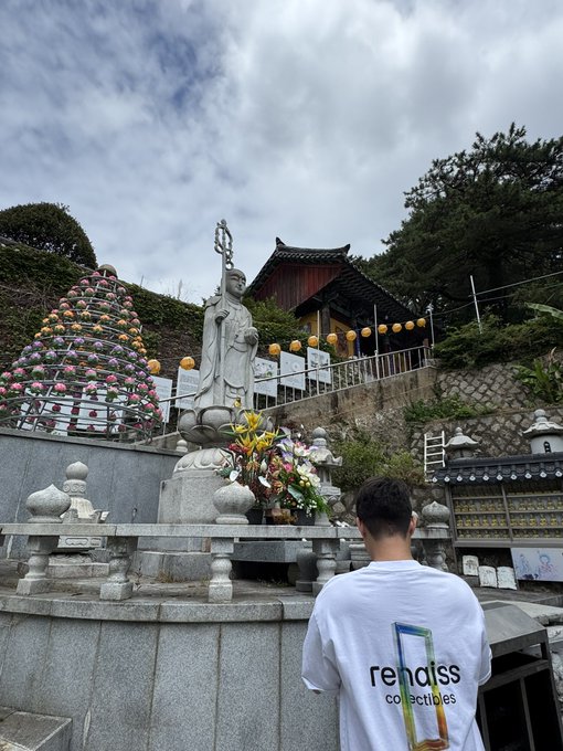 A person wearing a white t-shirt reading 'rendiss collectives' stands before a Buddhist statue on a lotus pedestal at what appears to be a temple, with a tree decorated with colorful ornaments in the background.