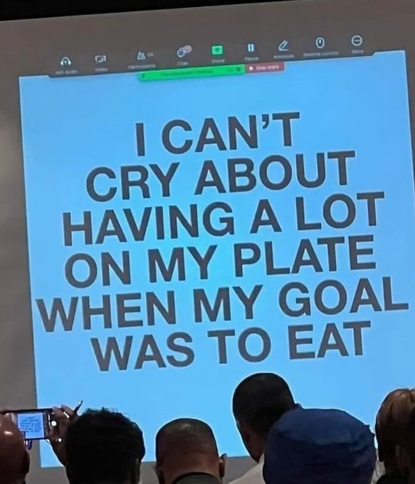 A presentation slide displayed at a meeting reads, "I CAN'T CRY ABOUT HAVING A LOT ON MY PLATE WHEN MY GOAL WAS TO EAT," as audience members look on, some recording the moment on their phones.
