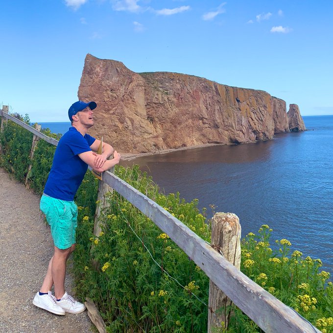 A man in a blue shirt, turquoise shorts, and a baseball cap leans on a wooden fence, gazing out at a large coastal rock formation and the ocean under a clear sky.