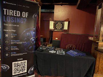 The image shows a table with various items, including a dartboard, a sign promoting a game or event, and some books. The background features a red wall and a wooden door. A banner can be seen hanging above the table, which reads "Tired of Losing? This win's on us!: How to Play: [0.17, 0.22, 0.5, 0.53]".