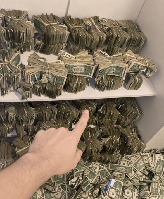 A hand points at shelves stacked with bundles and piles of US one-dollar bills inside what appears to be a large storage container.