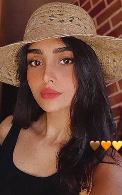 A young woman with long dark hair and red lipstick wears a straw sun hat and a black top, posing for a selfie against a brick wall with three yellow heart emojis visible in the corner.
