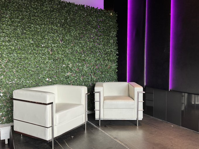 Two white leather armchairs with chrome frames sit on a stage in front of a lush green hedge wall, with purple lighting illuminating the adjacent black wall—creating a sleek, modern setting for the live podcast interviews hosted by @ImChrisChampion.