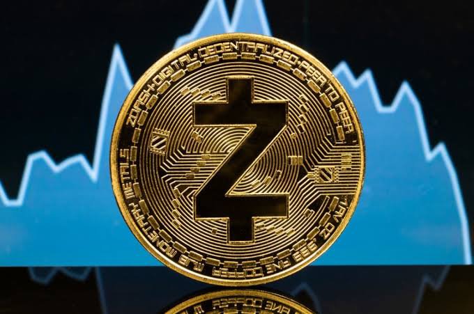 A physical gold-colored Zcash (ZEC) cryptocurrency coin displayed in front of a blue line chart showing an upward trend.
