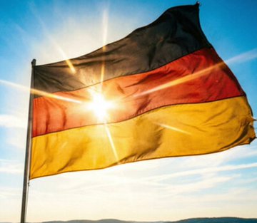 The German flag waving against a blue sky with the sun shining brightly behind it.