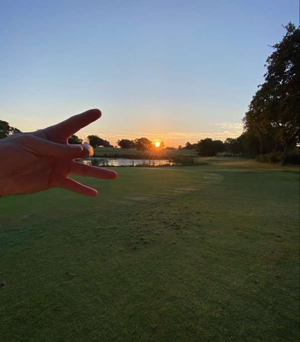 A hand reaches out toward a sunset over a golf course, with the sun positioned between the thumb and fingers in a forced perspective trick, with a pond and trees visible in the background.
