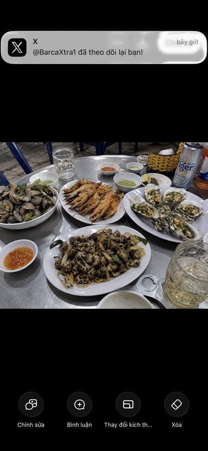 Screenshot of an X post by @BarcaXtra1 saying 'đã theo dõi lại bạn!' above a photo of a seafood feast (shrimp, oysters, snails, clams) on a metal table.