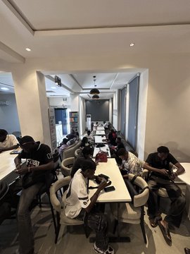A photograph of a group of people sitting along a long white conference table in a modern office space, with several individuals wearing face masks and looking at their phones or resting.