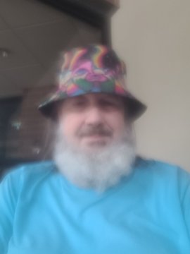 A selfie of a bearded older man wearing a colorful psychedelic bucket hat and a blue shirt.