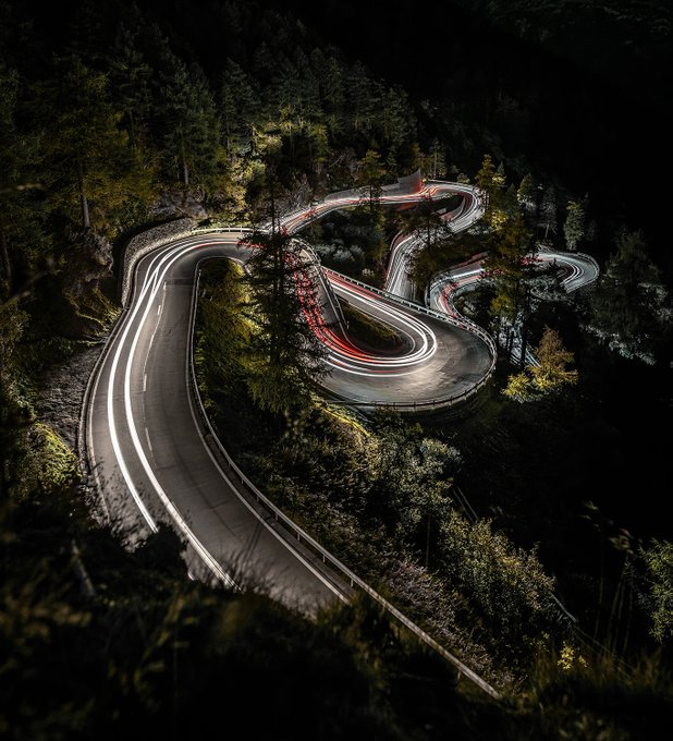 A striking long-exposure aerial photograph of a winding mountain road at night, with glowing white and red light trails from vehicles creating ribbon-like curves through a dark forested landscape.