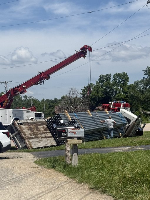 A red crane is lifting a load from a truck carrying large dark pipes that appears to have tipped over on a roadside, with a man standing nearby and a fire truck visible in the background.
