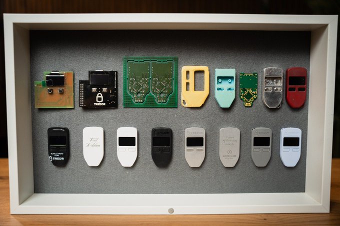 A framed display showcases the evolution of Trezor hardware wallets, featuring circuit boards alongside various device casings, including one labeled "9" and another marked "Bitcoin." The collection illustrates Trezor's hardware development, reinforcing the article's message about the importance of secure, self-custody tools in maintaining Bitcoin's original vision of financial sovereignty.