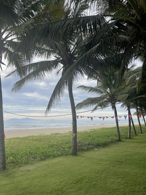 A tropical beach scene with palm trees, green grass, colorful bunting flags strung across, and the ocean under a cloudy sky.