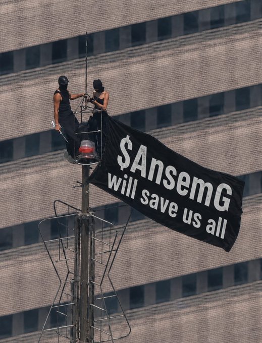 Two activists on top of a light pole unfurling a black banner reading '$AnsemG will save us all' against a city building backdrop.
