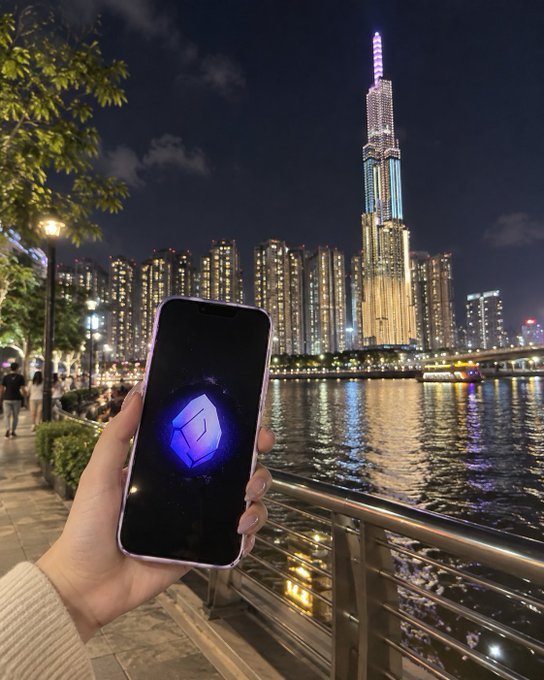 A hand holds a smartphone displaying a glowing blue crypto logo against the backdrop of a brightly lit skyscraper and waterfront city skyline at night.