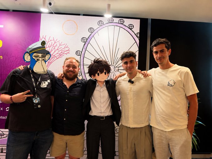Five men pose together indoors, with two having their faces obscured by cartoon avatars (a blue ape with sunglasses and an anime character), standing in front of a wall graphic featuring a ferris wheel design.