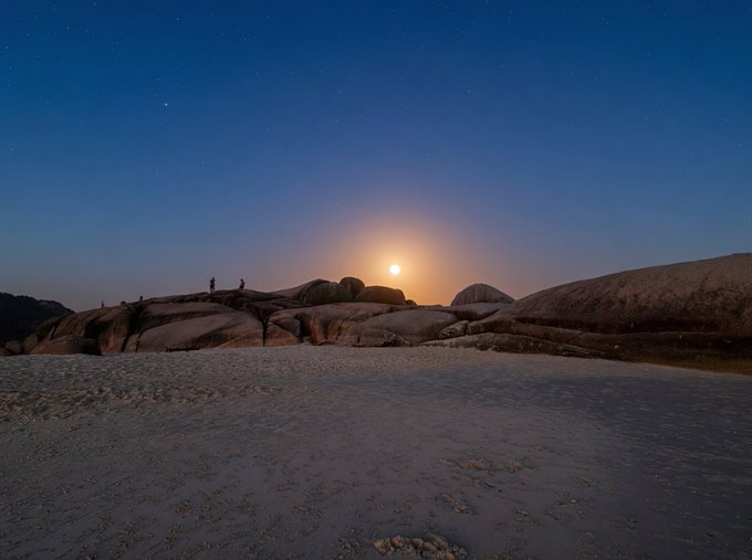 A moonlit beach scene at night with a glowing moon low on the horizon casting warm light over smooth rocks and sand, where tiny silhouettes of people can be seen standing on the rocky outcrop, perfectly capturing the serene "Midnight Glow" atmosphere described in the post.