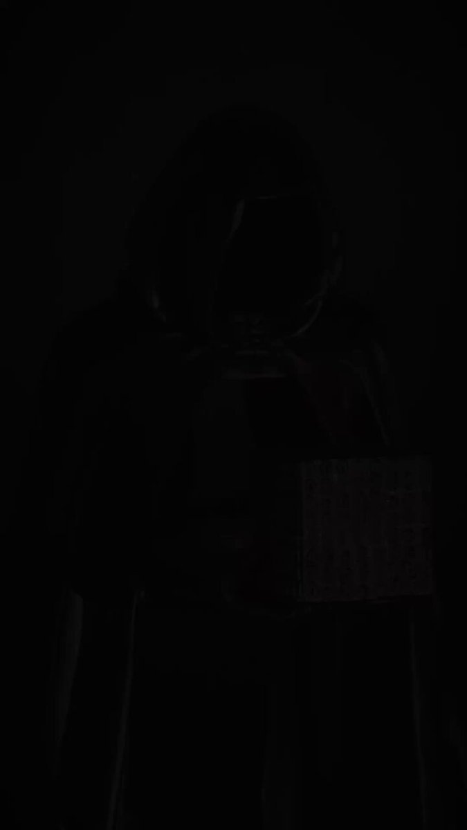 The image shows a person wearing a hoodie standing against a dark background with their face obscured by shadow. The person's position, along with the darkness of the scene, creates an atmosphere that suggests they are hiding or blending into the surroundings. The caption provided seems unrelated and does not provide any context about the image or its content.