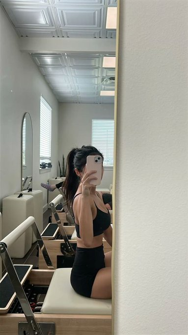 A woman takes a mirror selfie at a Pilates studio on her reformer machine, wearing a black sports bra and shorts as she works out on a Friday afternoon—a deliberate break from her 24/7 crypto and AI trading routines to invest in her physical health alongside her long-term portfolio.