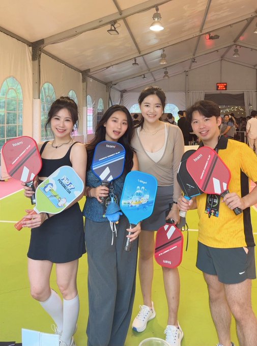 Four young adults pose together at a tented indoor venue, each holding colorful 'taarni' pickleball paddles.