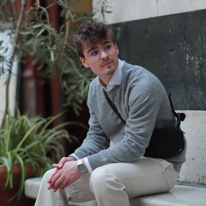 A young man with glasses sits thoughtfully on a stone bench, wearing a gray sweater over a white shirt and carrying a crossbody bag. The image complements the caption by visually capturing the introspective, milestone moment of leaving a corporate career to pursue full-time work in the Solana ecosystem.