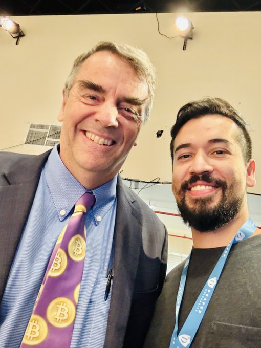 A selfie of two men smiling, with the older man on the left wearing a purple tie featuring Bitcoin logos, and the younger man on the right wearing a lanyard.