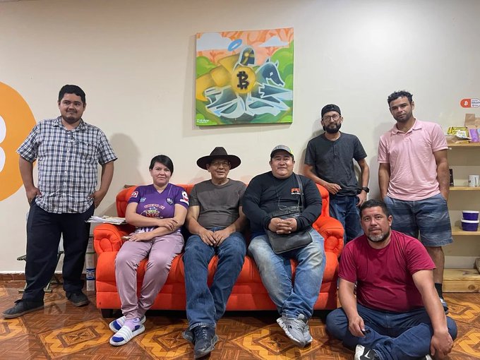 Six people pose together in front of a bright orange couch beneath a colorful graffiti-style Bitcoin painting.