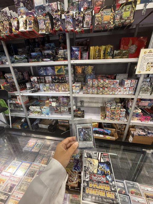 Inside a trading card store, a person's hand holds a card in front of shelves packed with merchandise; a "Yu-Gi-Oh! 2-PLAYER STARTER SET" box is visible on the glass counter.