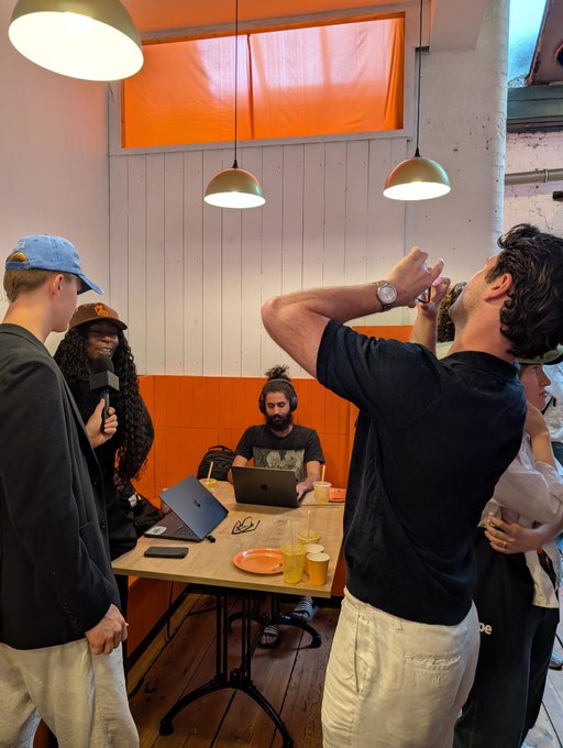 A group of people gather in an orange-walled café with pendant lights, where one man stands and drinks while another sits at a table with two laptops.