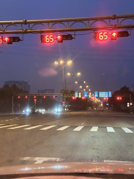 A driver's perspective at an intersection at dusk shows red traffic signal lights displaying "65" with upward arrows, while streetlights illuminate the multi-lane road ahead.