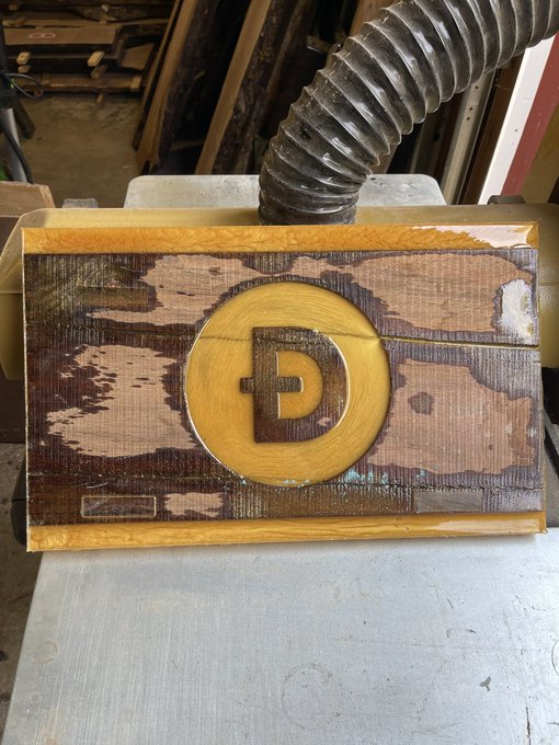 A wooden sign featuring a Dogecoin (DOGE) logo being finished on a workbench with a dust collection hose visible in the background.