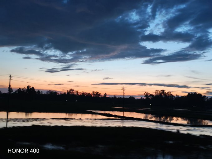 A sunset landscape taken with HONOR 400 phone, showing silhouettes of trees and power lines against a colorful sky reflected in water below.