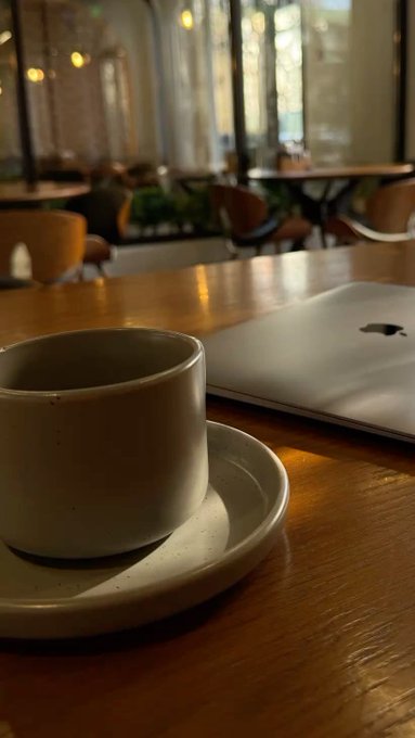 A white ceramic coffee cup and saucer sit on a wooden café table next to a silver Apple laptop, with large windows and warm lighting visible in the blurred background.