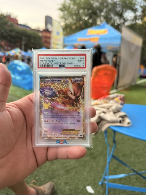 A hand holding a PSA-graded 2021 Pokemon Celebrations Mewtwo EX holographic trading card (#54, Mint 9) at what appears to be an outdoor event with blue tents.