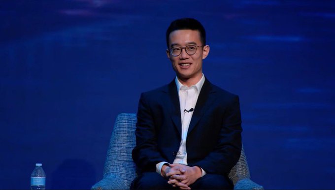 A man in a dark suit and glasses smiling while seated on a chair on stage against a blue backdrop, with a water bottle visible beside him.