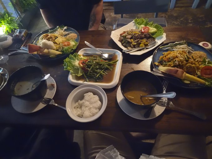 A table full of various Thai dishes including rice, curries, stir-fried vegetables, and salads, with a person's torso visible at the top.