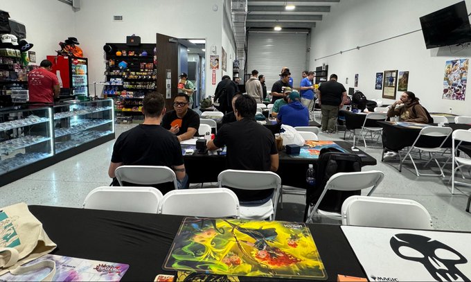A bustling trading card and hobby store, with customers seated at long tables playing games like Magic and Yu-Gi-Oh!, with shelves of merchandise in the background.