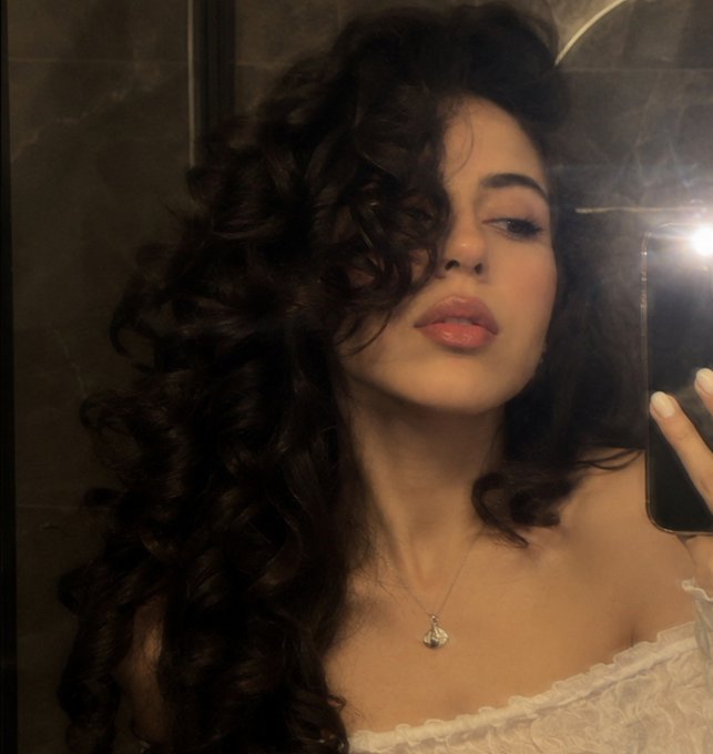 A woman with long, curly dark hair takes a mirror selfie, wearing a white ruffled top and a silver necklace, with the phone flash reflecting in the mirror.