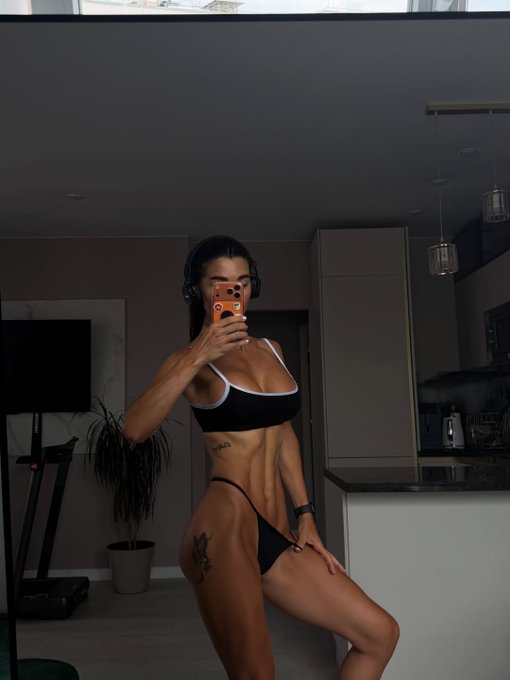 A fit woman wearing a black sports bra and bikini bottoms takes a mirror selfie with an orange phone in a modern apartment, showing visible abdominal and thigh tattoos.