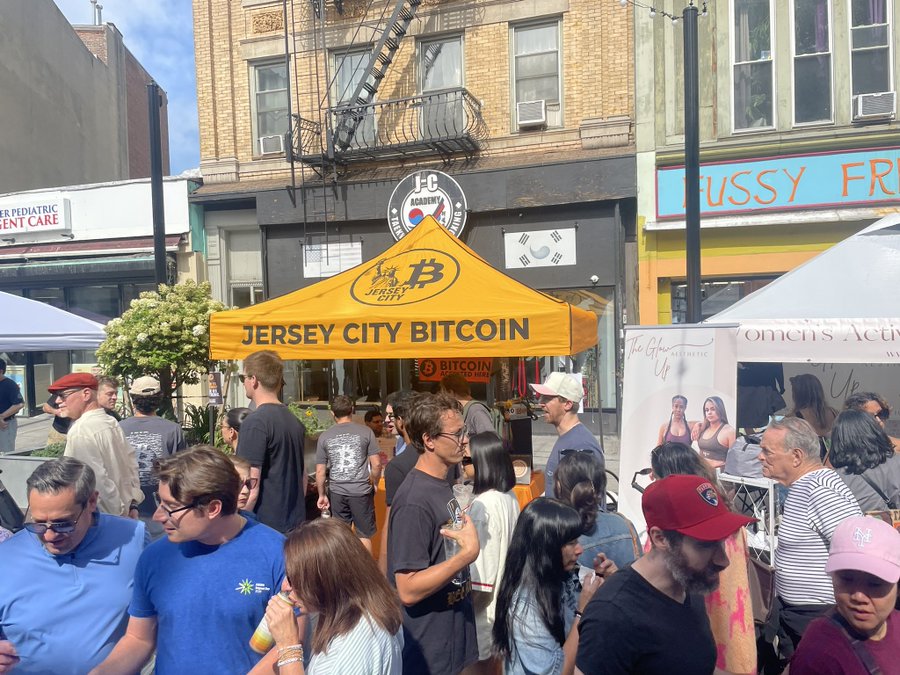 A bustling street fair features a vibrant orange tent labeled "JERSEY CITY BITCOIN" with a "Bitcoin Knowledge Center" sign, attracting a diverse crowd of attendees exploring financial education about cryptocurrency. The image aligns with the post by showcasing the Bitcoin educational booth as a key attraction at the Jersey City street fair, with nearby storefronts including a "Pussy Foot" shop and "Women's Aesthetic" booth adding to the lively community atmosphere.