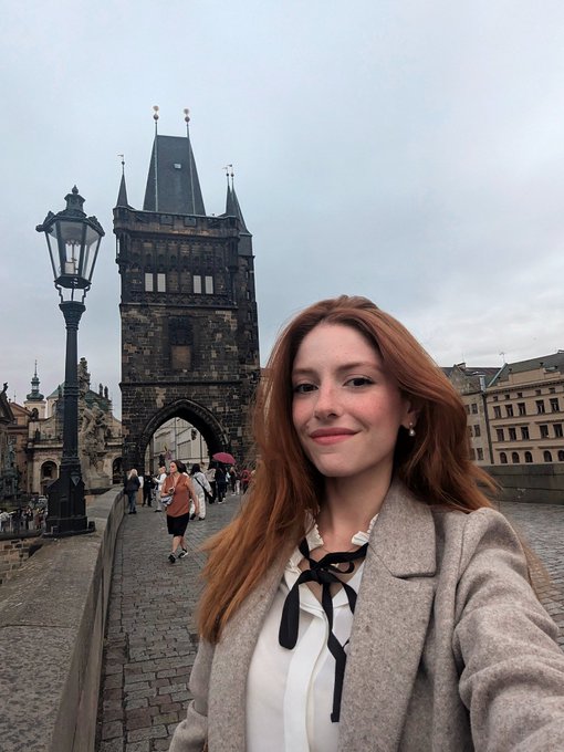 The image features a woman wearing a white shirt, black bow, and beige coat standing next to an old stone bridge over water. The photo has been edited to include a caption that reads "Bitcoin is the only money with zero new deposits to discover and control." This adds context to the scene by suggesting that the woman might be interested in Bitcoin or cryptocurrency, given its association with digital currency and the idea of discovering something new.