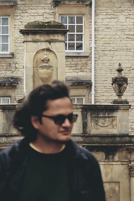 A man wearing sunglasses standing in front of an ornate historic stone building with decorative urns and carved architectural details.
