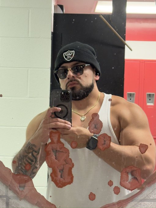 A mirror selfie of a muscular man wearing a Raiders beanie, sunglasses, and a white tank top, with red lipstick kiss marks overlaid on the photo.