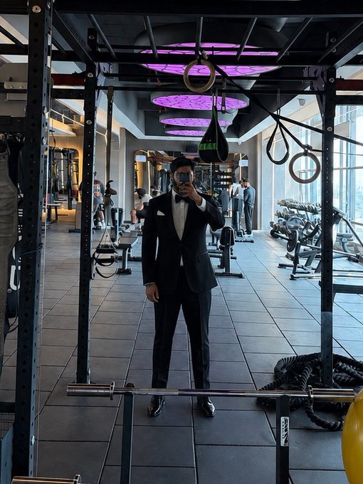 A man in a formal black tuxedo with a bow tie takes a mirror selfie inside a fully equipped gym, complete with squat racks, battle ropes, dumbbells, and gymnastic rings. The humorous juxtaposition of his formal attire against the hardcore fitness environment perfectly captures the irony the post seems to celebrate—someone truly "gets it."