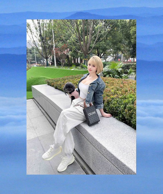 A young woman with blonde hair sits on a stone bench in a park-like setting, holding a small black dog and a Celine shopping bag, wearing a denim jacket and white outfit.