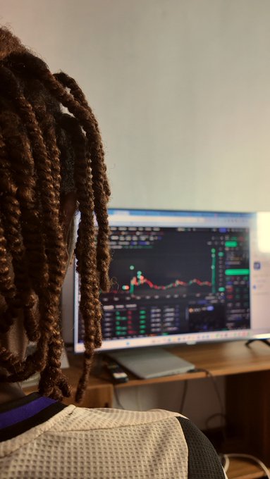 An over-the-shoulder view shows a person with braided hair wearing a mesh chair back, looking at a computer monitor displaying a financial trading chart with candlestick patterns.