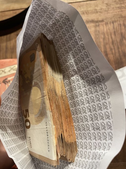 A hand holds a partially opened paper sleeve containing a thick stack of 50 Euro bills, with the serial number Z2206922605 visible, tied to a post referencing a Solana ($SOL) address.