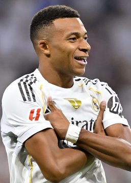 Soccer player Kylian Mbappé in a white Real Madrid jersey with 'Emirates' sponsorship, smiling with his hand on his chest.