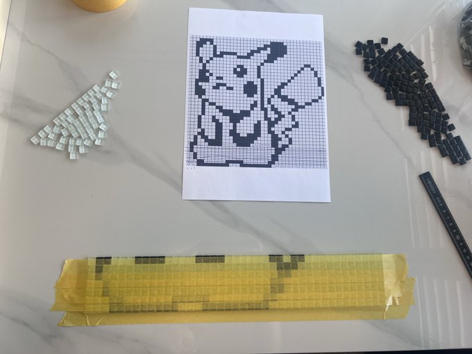 A Pikachu pixel art pattern printed on graph paper, surrounded by loose white and black fuse beads and a metal ruler on a marble surface.